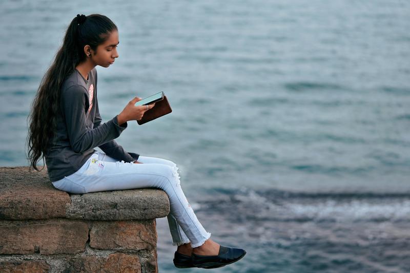 Image of a young woman sitting near the sea using a mobile phone