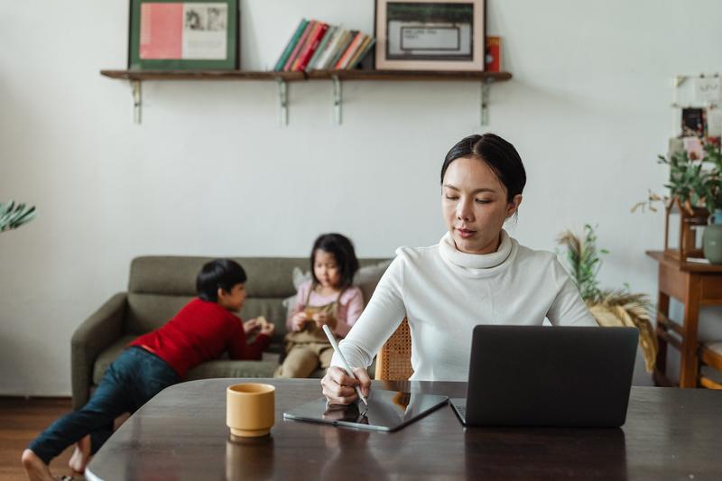 Young mother works remotely while children play on sofa behind