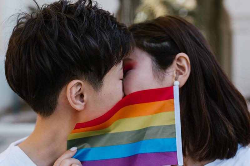 Two women kiss behind a LGBTQ+ pride flag
