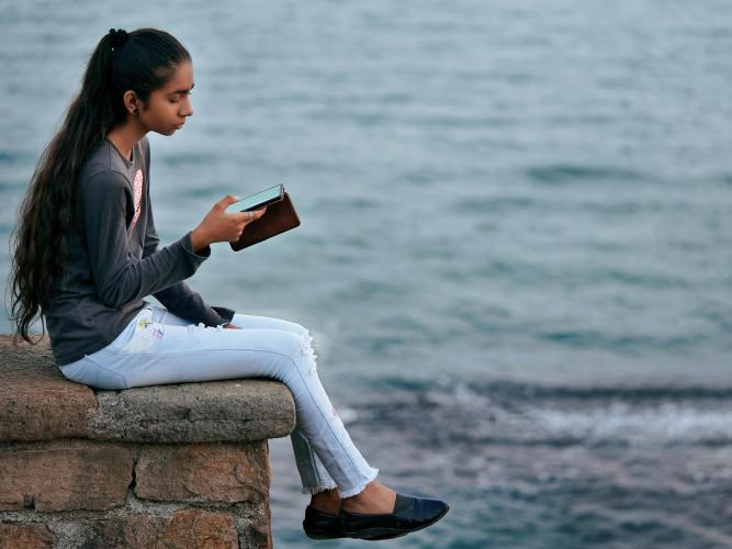 Image of a young woman sitting near the sea using a mobile phone