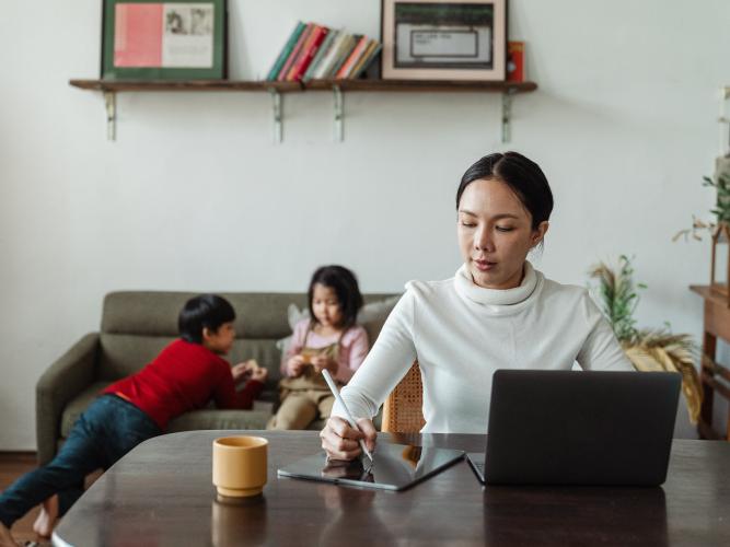Young mother works remotely while children play on sofa behind